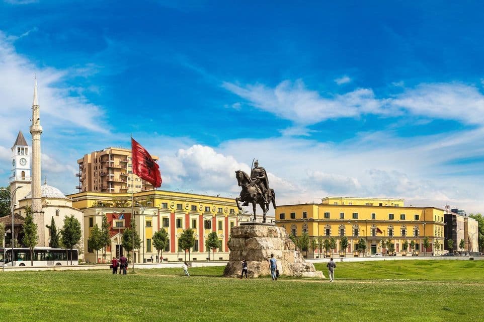 A bronze equestrian statue on a stone base in a grassy square, with a large Albanian flag and historic buildings in the background.