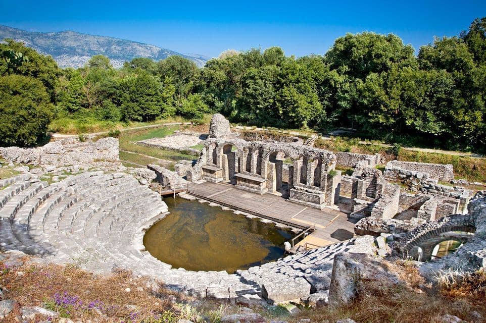 Les ruines d'un ancien amphithéâtre en pierre avec une fosse d'orchestre inondée, entourées d'une forêt dense et de montagnes lointaines sous un ciel bleu.