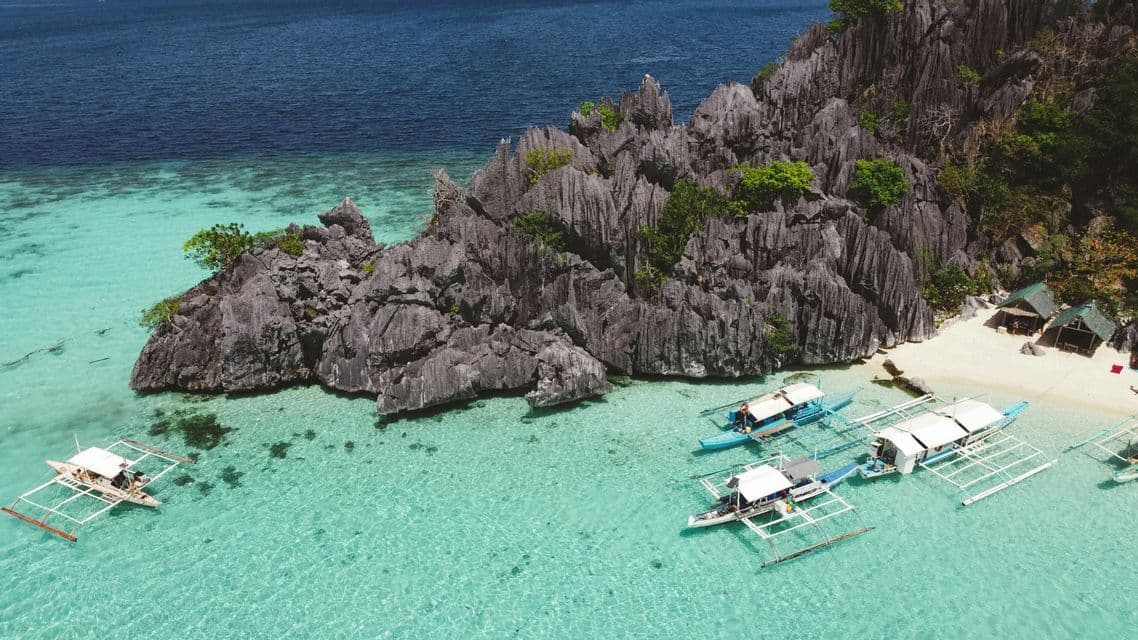 Luftaufnahme von Auslegerbooten, die in einer klaren türkisfarbenen Bucht neben einer zerklüfteten Felsformation und einem kleinen weißen Sandstrand ankern.