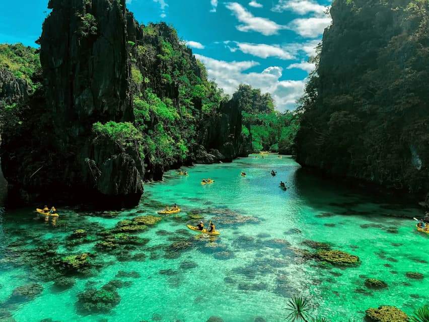 Eine WeRoad-Gruppenreise paddelt in gelben Kajaks durch eine klare türkisfarbene Lagune, umgeben von üppig grünen Klippen unter blauem Himmel.