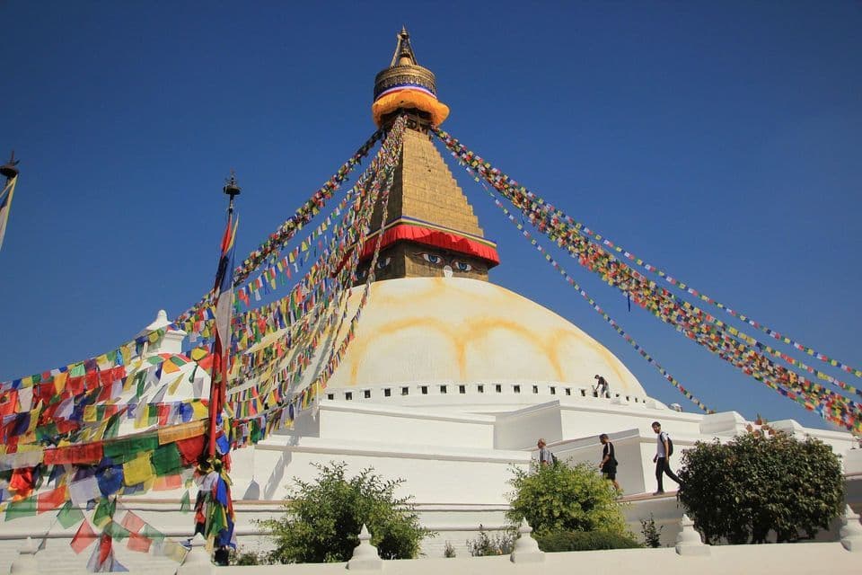 Eine weiße buddhistische Stupa mit goldener Spitze und aufgemalten Augen, davor wehen bunte Gebetsfahnen vor blauem Himmel.