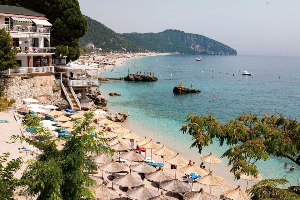 An elevated view of a crowded beach with many straw umbrellas, clear turquoise water, and a forested mountain coastline.