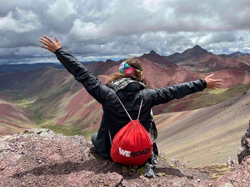 A traveler with a red WeRoad backpack sits on a rocky peak, arms outstretched, overlooking a colorful mountain range under a cloudy sky.