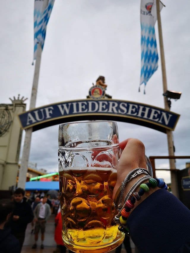 Una mano con un grande boccale di birra di vetro davanti a un'insegna ad arco con la scritta 'AUF WIEDERSEHEN' durante un festival.