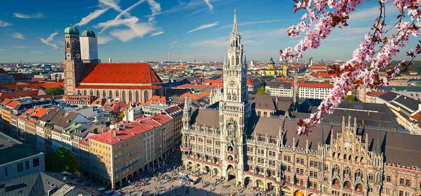 Una vista aerea di una storica città europea con architettura gotica e tetti in tegole rosse, incorniciata da fiori rosa