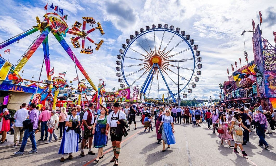 Una folla di persone, alcune in abiti tradizionali, cammina attraverso un luna park con una grande ruota panoramica e altre giostre.