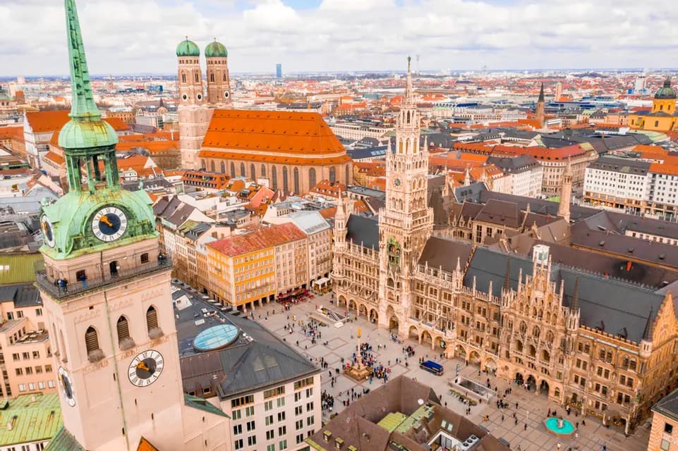 Una vista aerea di una storica piazza europea, che mostra una torre dell'orologio con una guglia verde e un edificio gotico ornato.