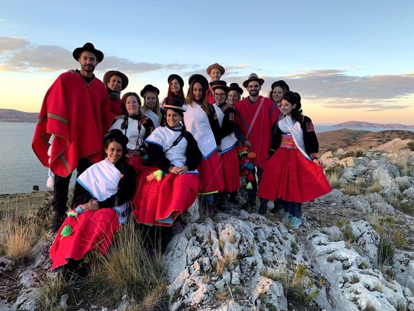 Un groupe WeRoad en voyage, vêtu de ponchos et jupes rouges traditionnels, pose pour une photo sur une colline rocheuse surplombant l'eau au coucher du soleil.