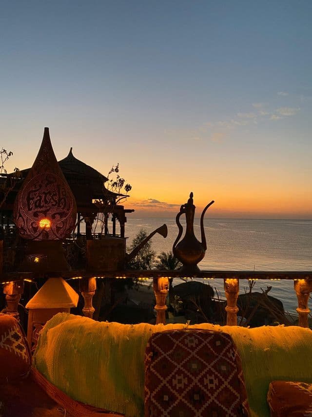 Una lanterna intarsiata e una brocca di metallo poggiano sul parapetto di un balcone, con vista sul mare al tramonto.