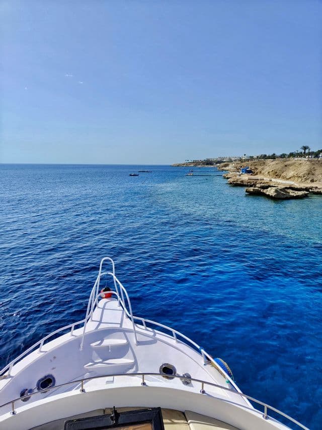 Una vista dalla prua di una barca bianca che naviga su acqua blu profondo lungo una costa rocciosa sotto un cielo sereno.