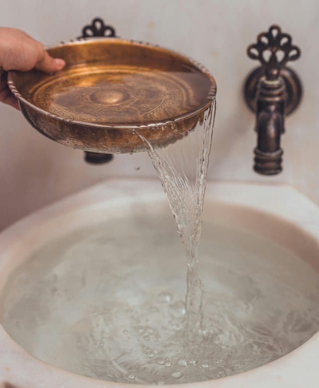 Una mano versa acqua da una brocca d'ottone ornata in un lavabo di marmo bianco con un rubinetto vintage sullo sfondo.