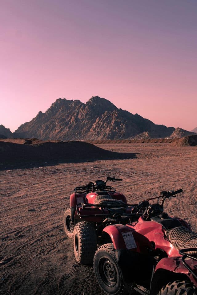 Due quad rossi parcheggiati su terreno desertico sabbioso, con una montagna rocciosa sullo sfondo sotto un cielo rosa.