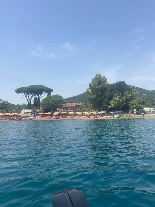 Vista da una barca sull'acqua blu, guardando verso una spiaggia con ombrelloni gialli e alberi contro una collina.