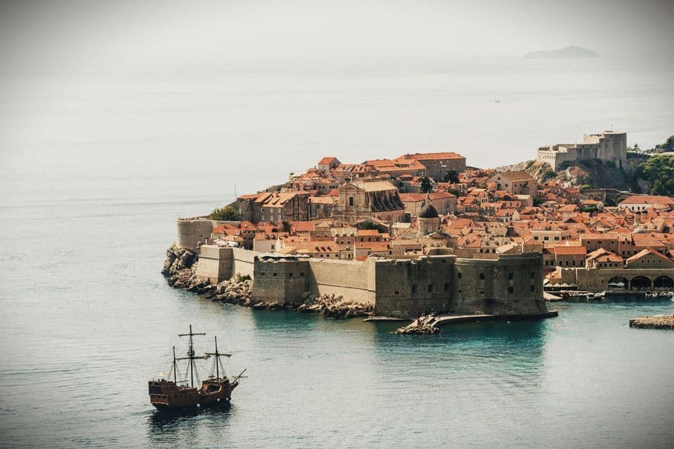 Una nave di legno d'epoca naviga su acque turchesi di fronte a una storica città murata con tetti di tegole arancioni.