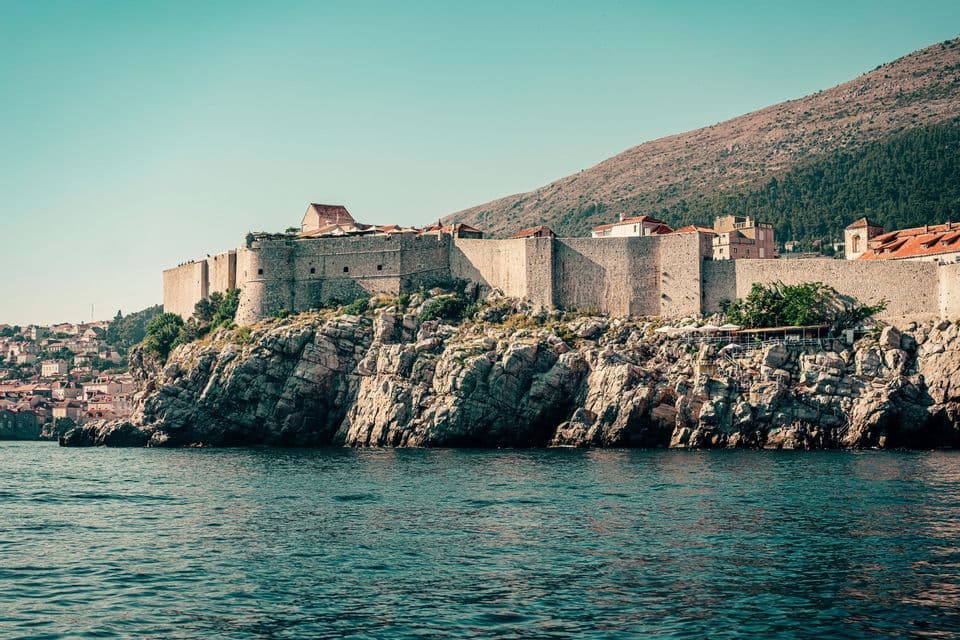 Una vista dall'acqua di una grande fortezza in pietra costruita su una scogliera rocciosa, con una collina sullo sfondo.