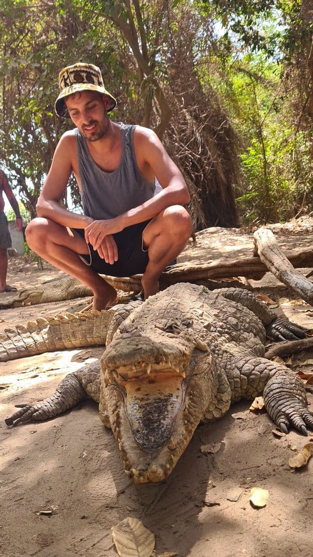 Ein Mann einer WeRoad Gruppenreise hockt im Sand hinter einem großen Krokodil mit weit geöffnetem Maul.