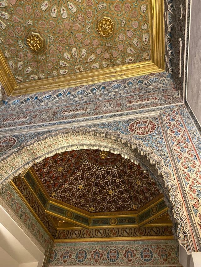 Un techo y un arco ornamentados cubiertos de intrincados patrones geométricos y caligráficos con detalles en oro, azul y rojo.