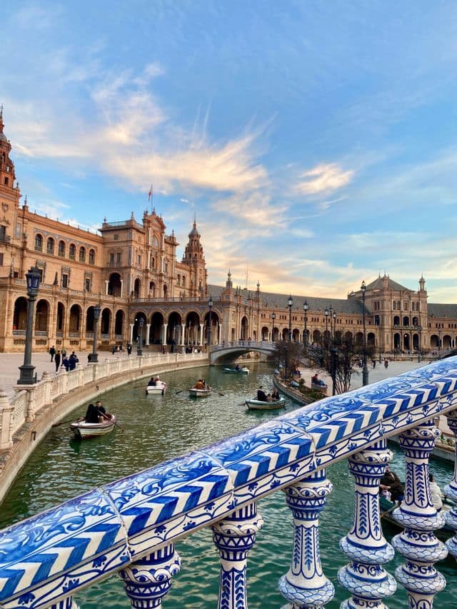 Una vista desde detrás de una barandilla de azulejos azules y blancos muestra a personas remando en botes en un canal frente a un gran edificio histórico.