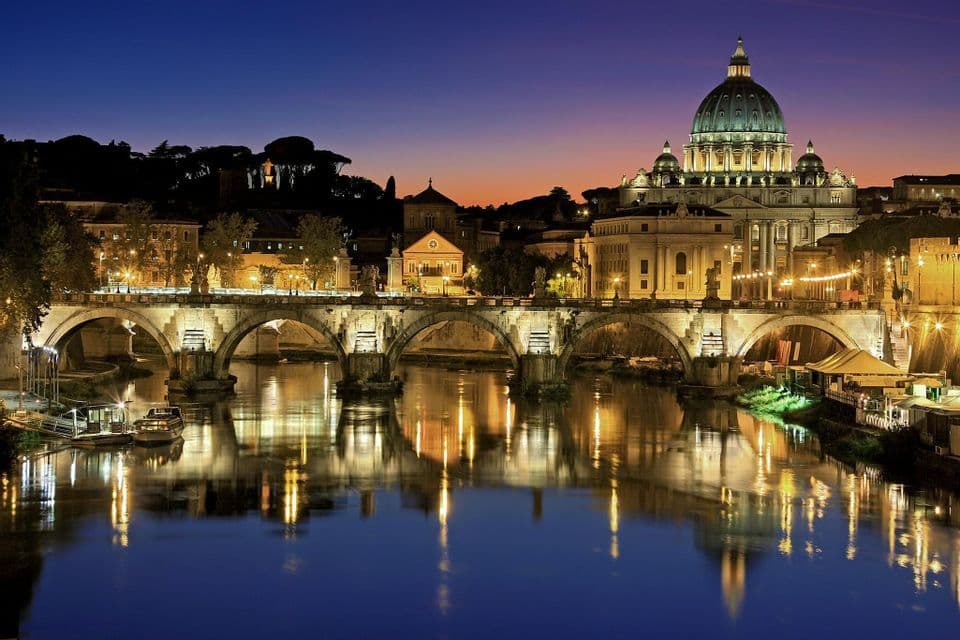 Un pont de pierre illuminé et une grande basilique à dôme se reflétant dans une rivière au crépuscule sous un ciel bleu foncé.