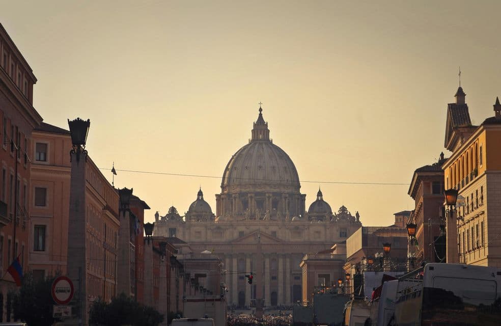 Una grande basilica a cupola alla fine di una strada, con folle in lontananza ed edifici su entrambi i lati, bagnata dalla calda luce del tramonto.