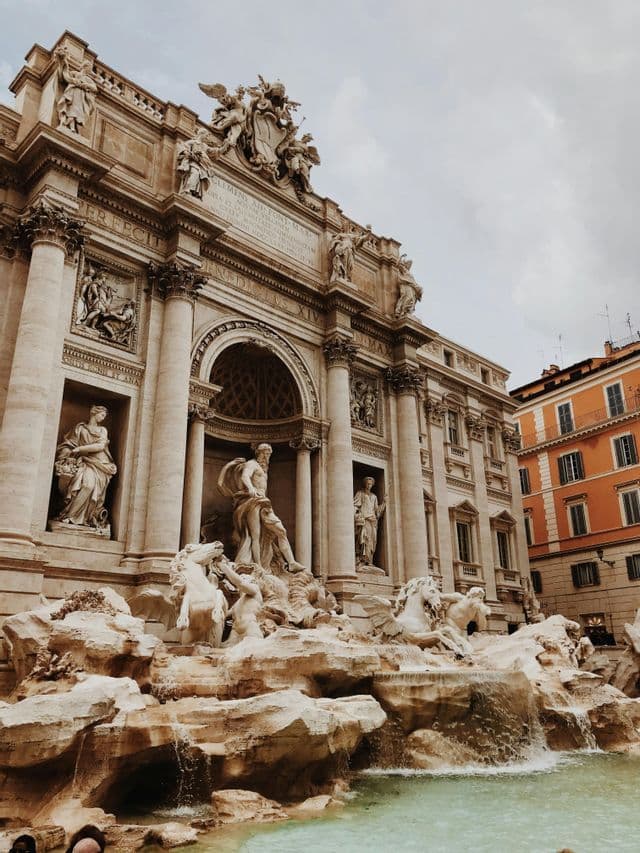 Un plano contrapicado de una ornamentada fuente barroca con grandes estatuas de figuras y caballos bajo un cielo nublado.