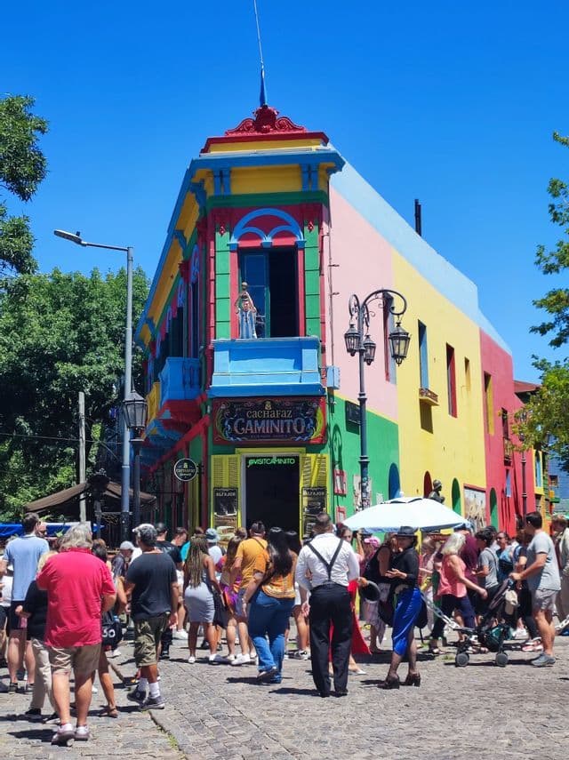 Una multitud de personas en una calle empedrada frente a un edificio de esquina de colores brillantes bajo un cielo azul claro.