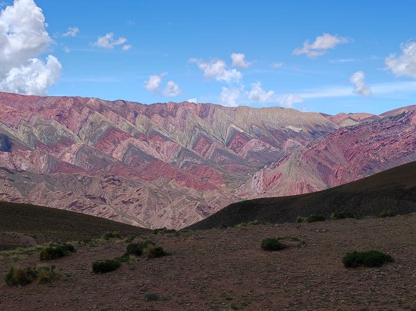 Una cordillera muestra capas vibrantes y multicolores de formaciones rocosas bajo un cielo azul con nubes dispersas.