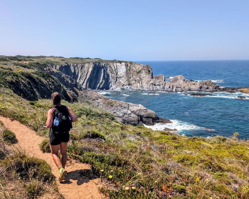 Eine Frau mit Rucksack rennt auf einem Feldweg entlang einer grasbewachsenen Klippe mit Blick auf eine felsige Küste und das Meer.