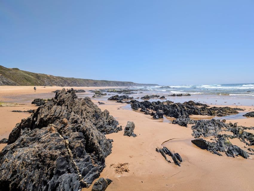 Dunkle, zerklüftete Felsen ragen aus dem nassen Sand eines breiten Strandes, mit Meereswellen und einer Person im Hintergrund unter einem klaren Himmel.