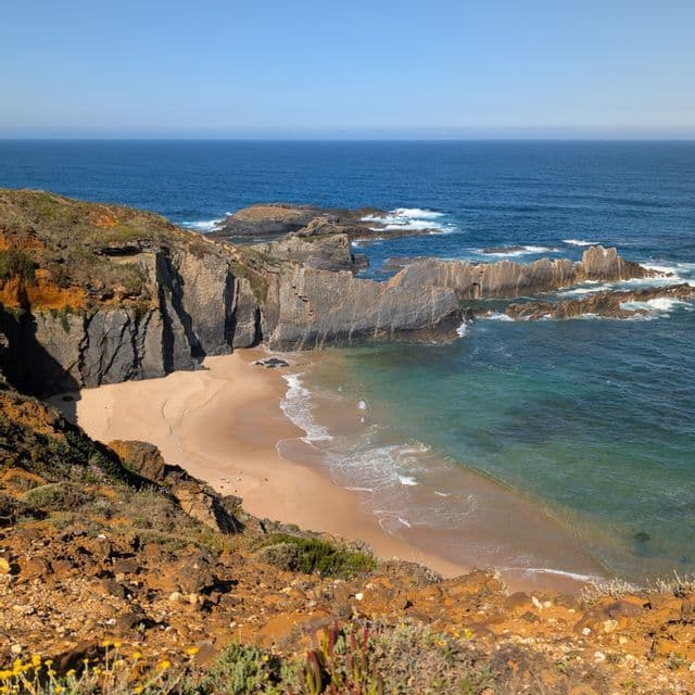 Luftaufnahme einer Sandbucht mit türkisfarbenem Wasser, flankiert von dunklen, geschichteten Felsenklippen und einem tiefblauen Meer.