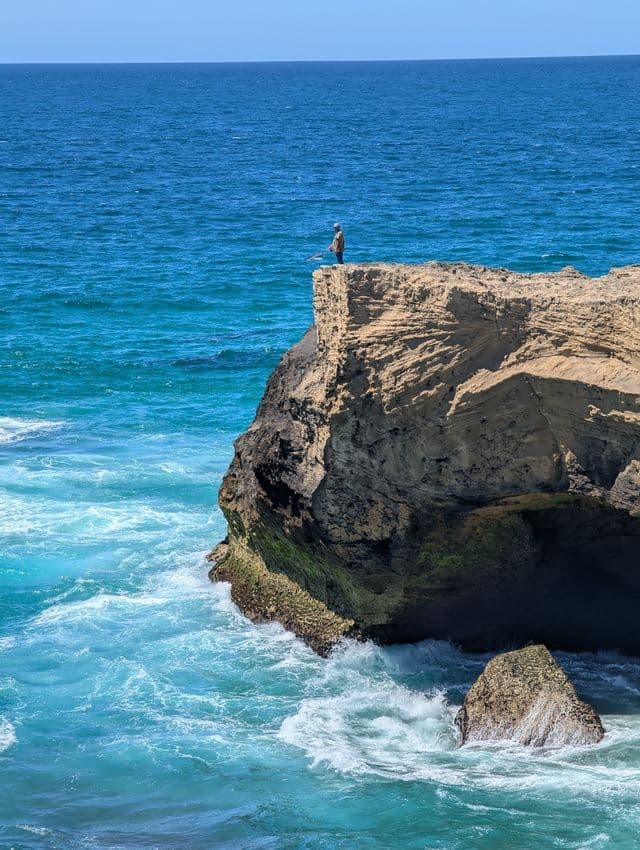 Eine Person mit Angelrute steht am Rande einer hohen Felsklippe und blickt auf einen türkisfarbenen und blauen Ozean.