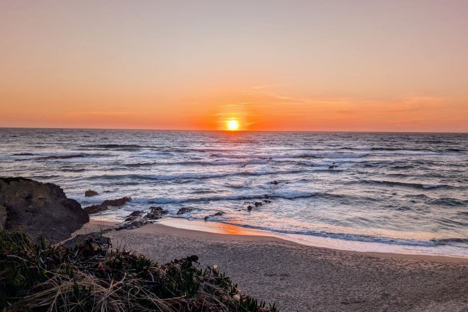Die Sonne geht über dem Meereshorizont unter und taucht die Wellen, die auf einen Sandstrand mit Felsformationen rollen, in ein oranges Leuchten.
