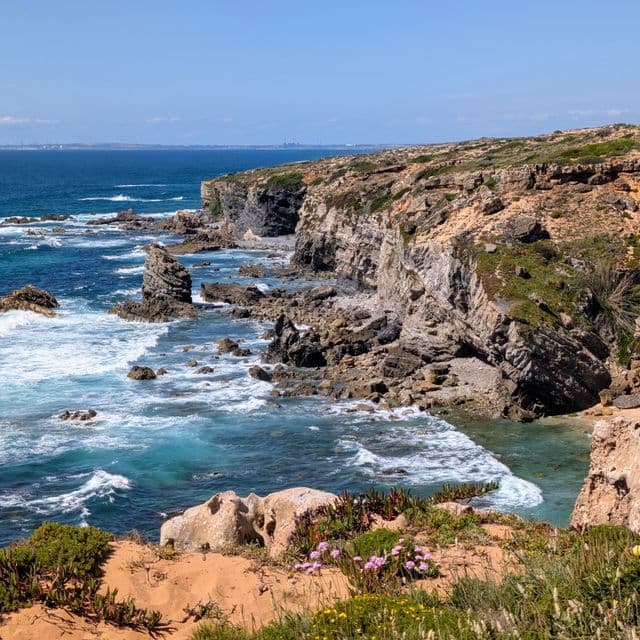 Eine zerklüftete Küste mit steilen Klippen und Brandungspfeilern, die unter einem klaren blauen Himmel von türkisfarbenen Wellen umspült wird.