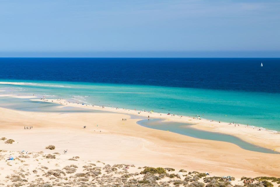Veduta dall'alto di una spiaggia sabbiosa, con persone sparse lungo la riva turchese e l'oceano blu profondo sullo sfondo.