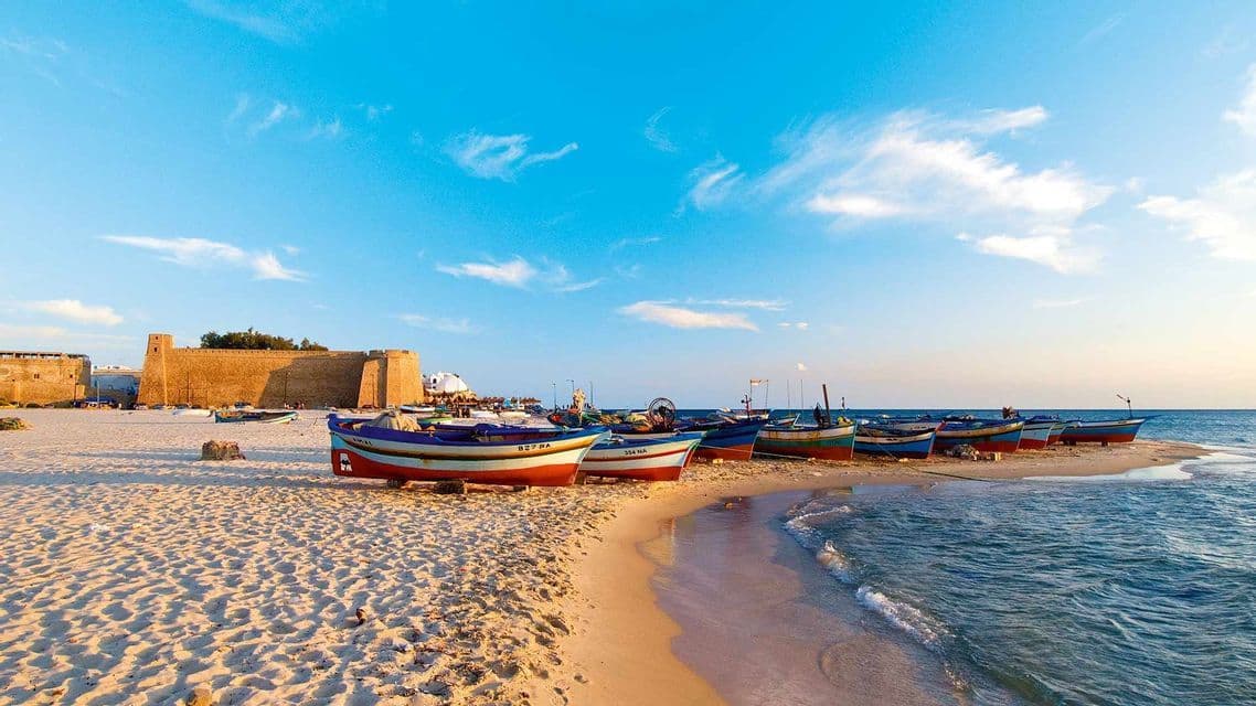 Une ligne de bateaux de pêche colorés repose sur une plage de sable au bord de la mer, avec une vieille forteresse en pierre à l'arrière-plan.