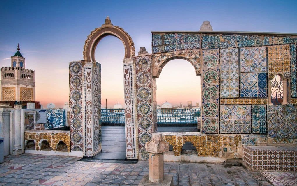 Une terrasse sur le toit avec des arches ornées et des mosaïques colorées surplombe un paysage urbain avec un minaret au coucher du soleil.