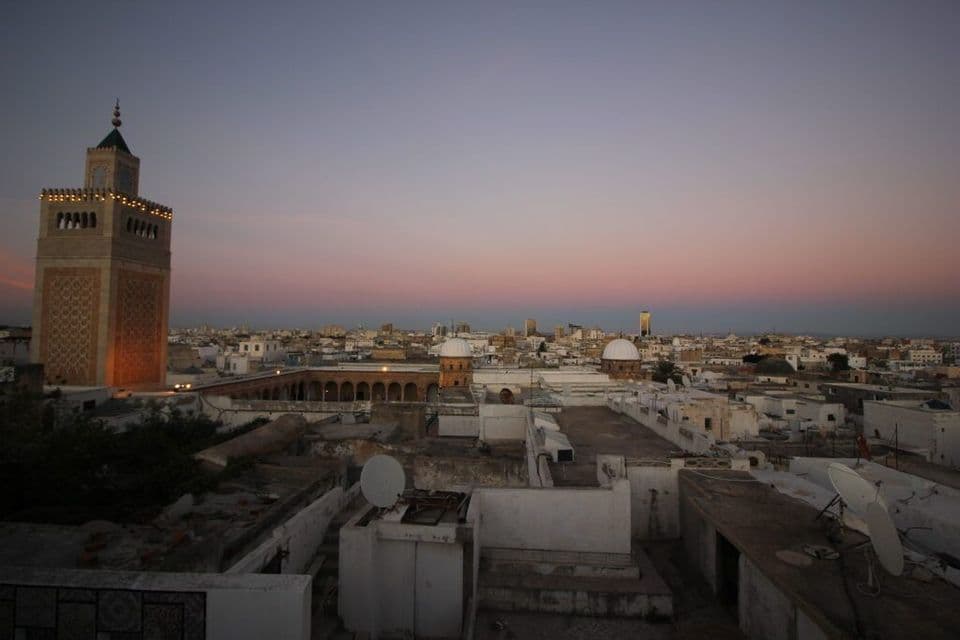Un minaret illuminé surplombe un paysage urbain de bâtiments blancs et de toits plats sous un ciel rose et violet au crépuscule.