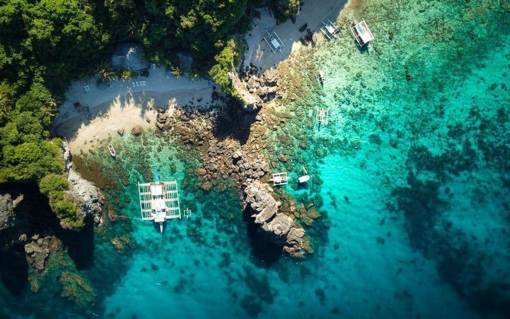 Una vista aerea dall'alto di una cala tropicale con una spiaggia di sabbia bianca, una foresta lussureggiante e barche ormeggiate in acque cristalline turchesi.