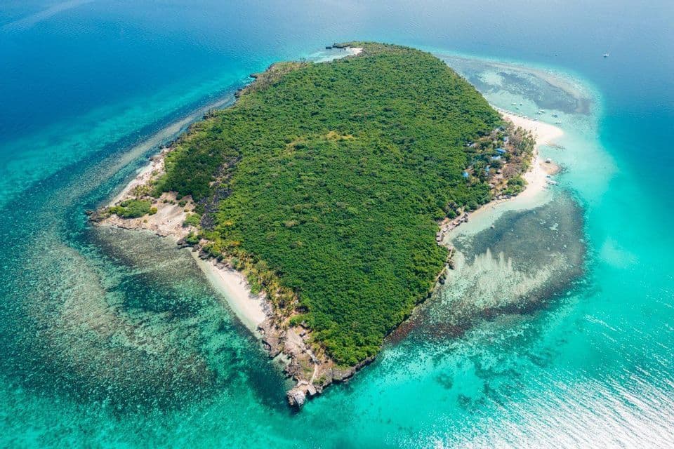 Una veduta aerea di un'isola tropicale coperta da una fitta foresta verde, circondata da acque turchesi e barriere coralline.