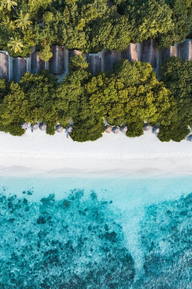 Vista aerea dall'alto di bungalow sulla spiaggia immersi in una foresta lussureggiante, a ridosso di una spiaggia di sabbia bianca e acque turchesi cristalline.