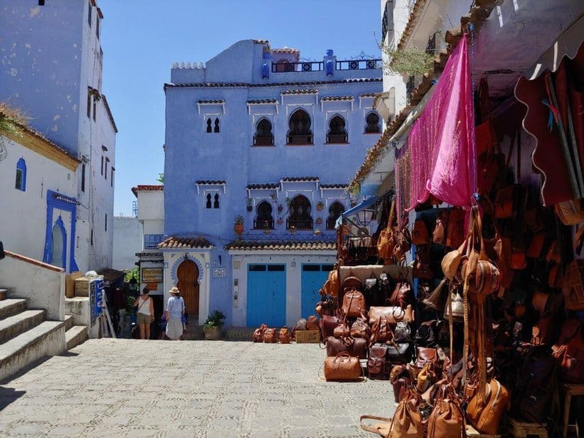 Auf einem Kopfsteinpflasterplatz vor einem großen, blauen Gebäude bietet ein Marktstand Ledertaschen zum Verkauf an.