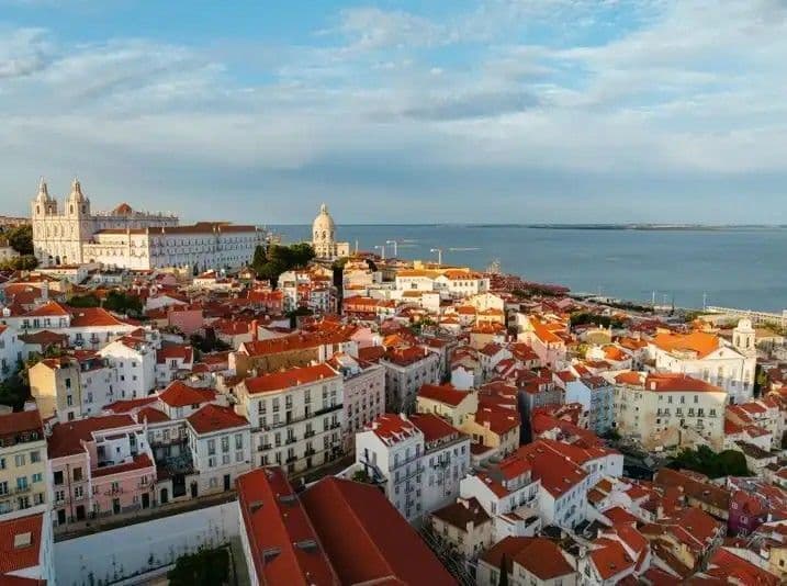 Eine Luftaufnahme einer Küstenstadt mit roten Ziegeldächern und historischen Gebäuden, die unter einem teilweise bewölkten Himmel auf das Wasser blicken.