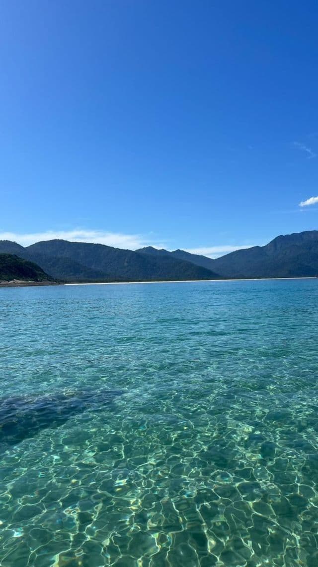 Le soleil se reflète sur des eaux turquoises claires et calmes, avec un littoral montagneux en arrière-plan sous un ciel bleu.