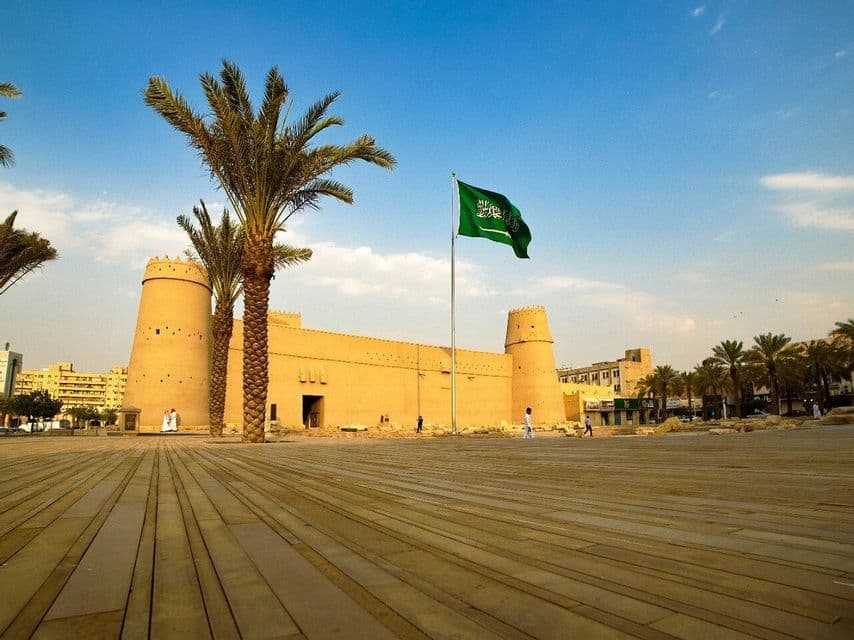 Le drapeau de l'Arabie Saoudite flotte devant une grande forteresse couleur sable, avec des palmiers et une large place.
