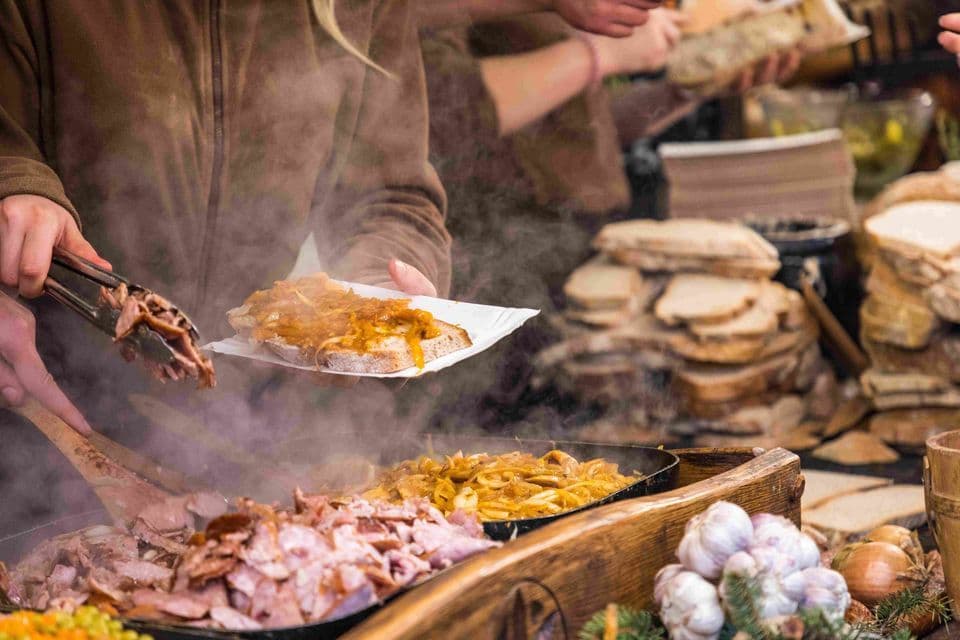 Un vendedor en un puesto de comida sirve carne y cebolla calientes de una sartén humeante sobre una rebanada de pan en un plato de papel.