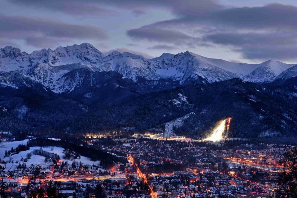 Un pueblo iluminado y una luminosa pista de esquí enclavados al pie de montañas nevadas, al anochecer y bajo un cielo nublado.