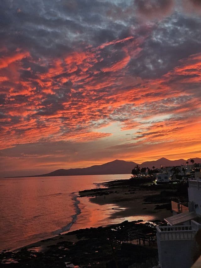 Una costa con edificios y una playa al atardecer, bajo un cielo lleno de nubes rojas y naranjas texturizadas sobre el mar.
