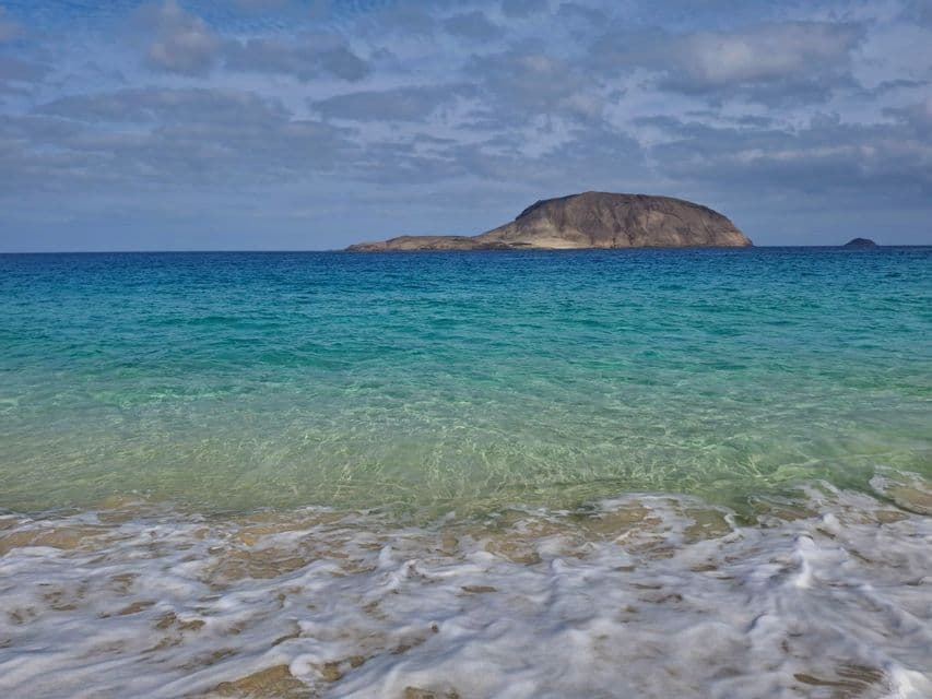 Una isla rocosa se asienta en agua oceánica clara y turquesa, vista desde una orilla arenosa con olas suaves y espuma blanca en primer plano.