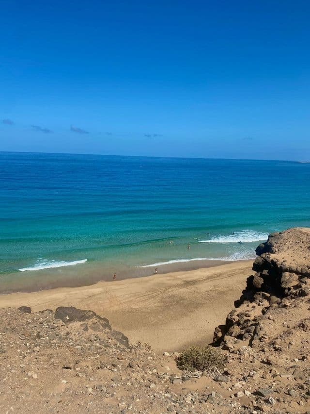 Una vista desde un acantilado rocoso con vistas a una playa de arena con gente nadando en el océano turquesa bajo un cielo azul claro.