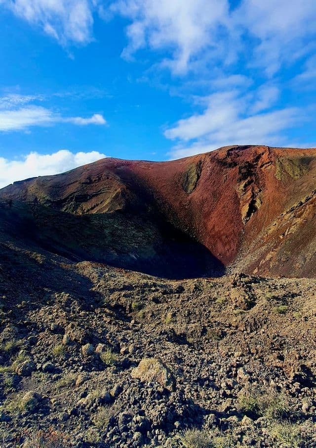 Un cráter volcánico de color marrón rojizo con laderas rocosas oscuras se asienta bajo un cielo azul parcialmente nublado.